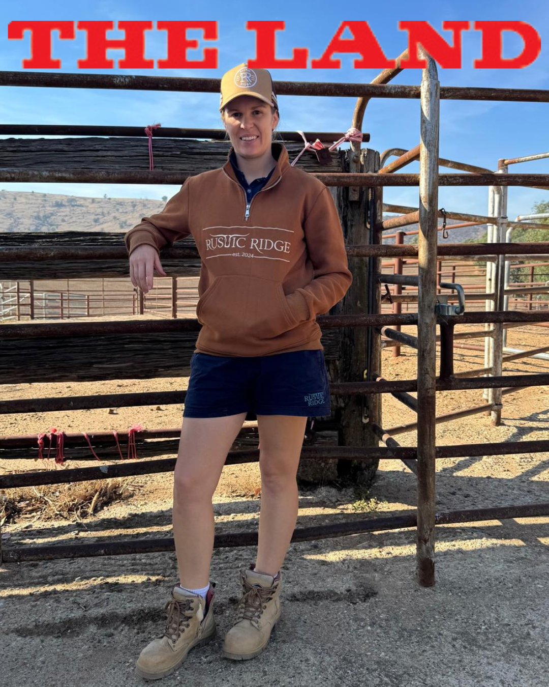 Marlee owner of Rustic Ridge leaning on a wooden cattle rail post in Rustic Ridge work shorts and 1/4 zip jumper.