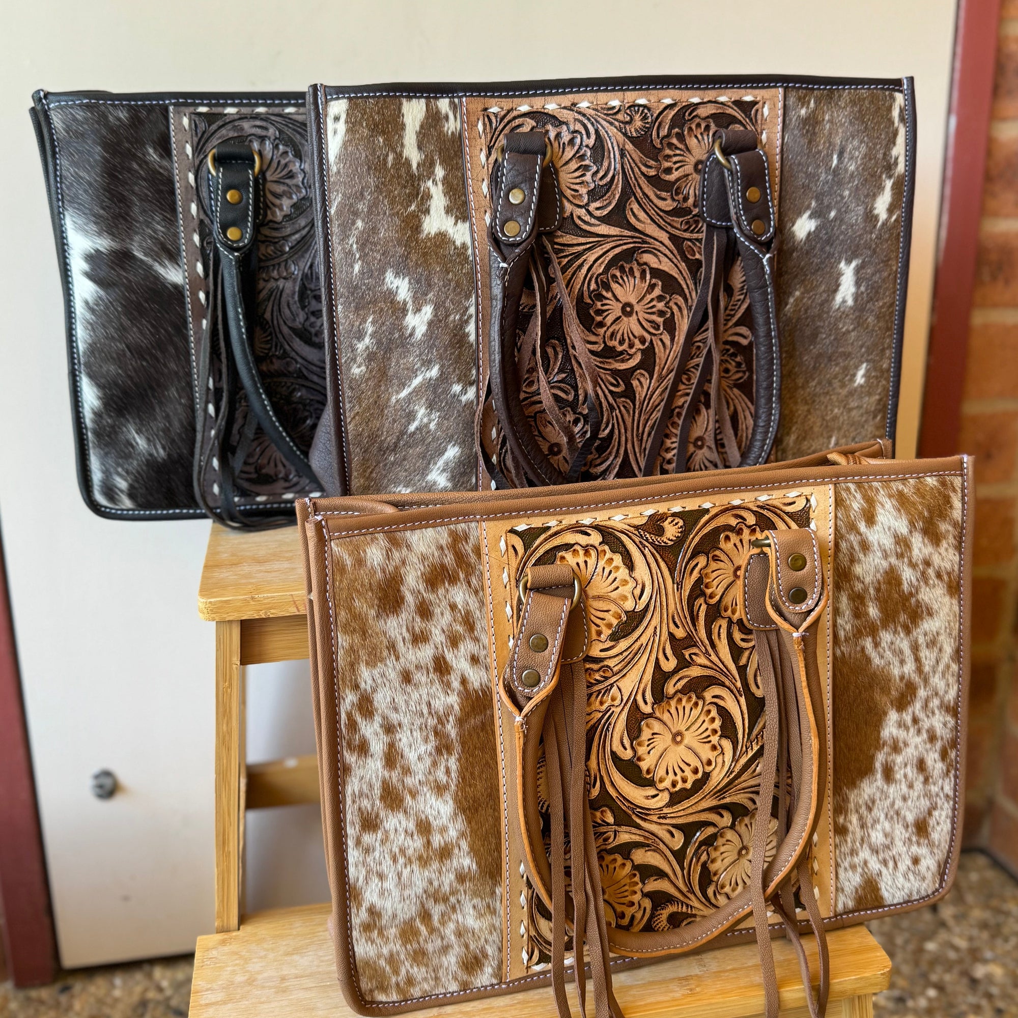 Three leather bags with intricate designs on a wooden stand against a brick wall.