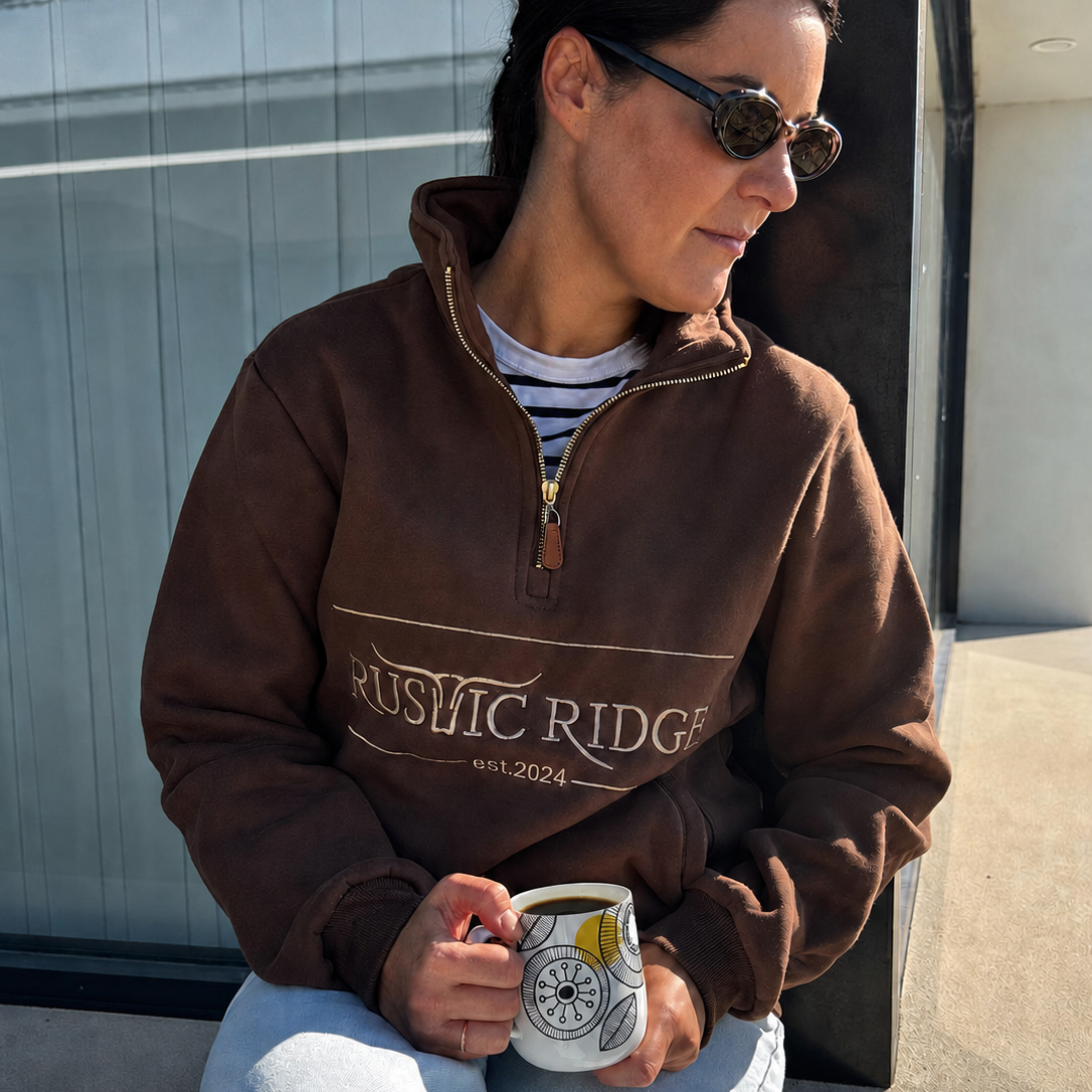 A woman wearing a brown quarter-zip sweatshirt with 'RUSTIC RIDGE est. 2024' printed on the front, sitting outdoors and holding a white mug.