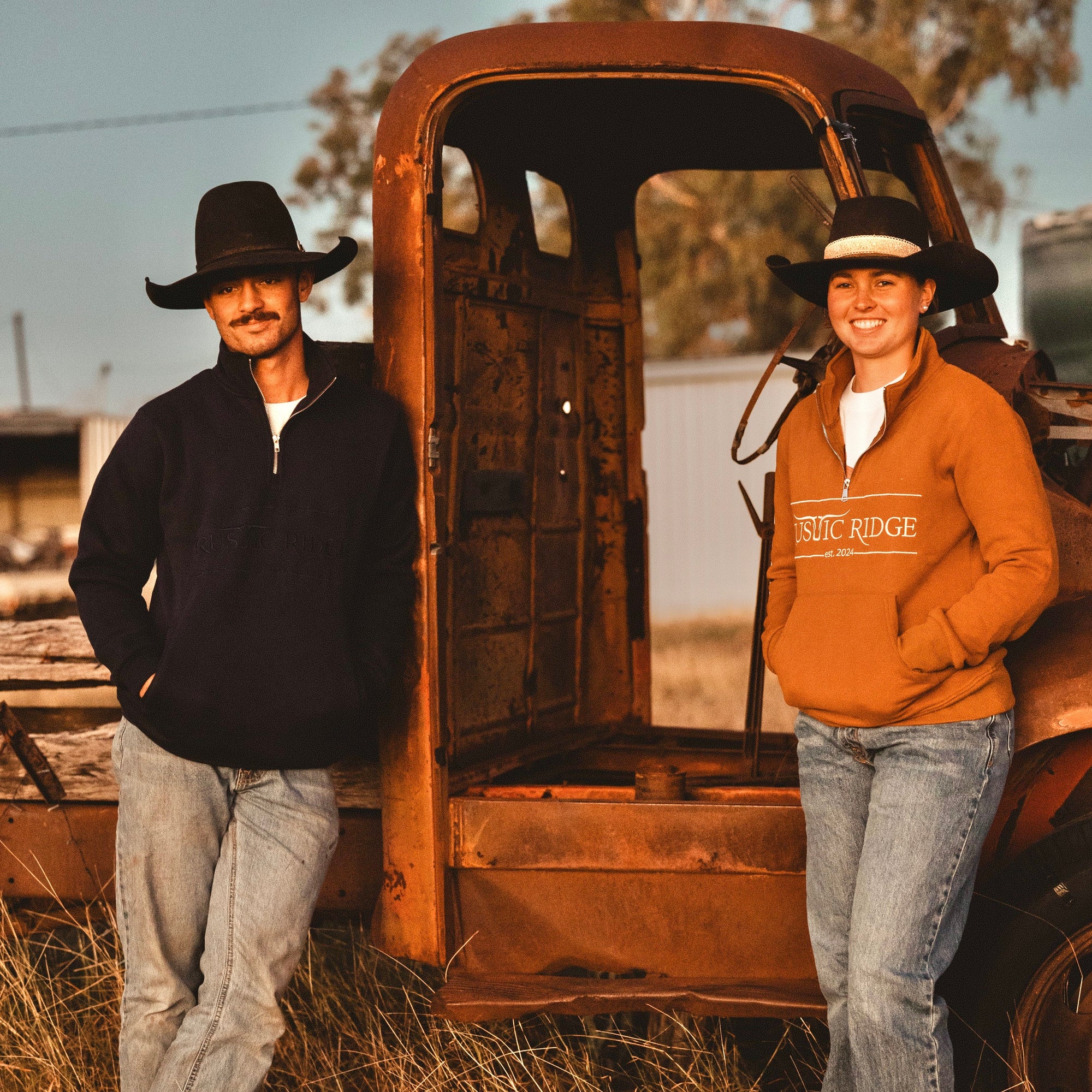 Two people standing next to an old truck in a field with a sunset sky.