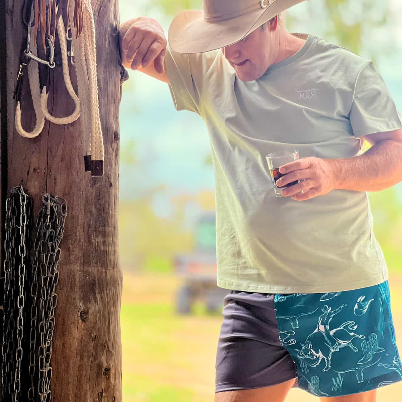 Man in a cowboy hat and casual clothing leaning against a wooden post outdoors.