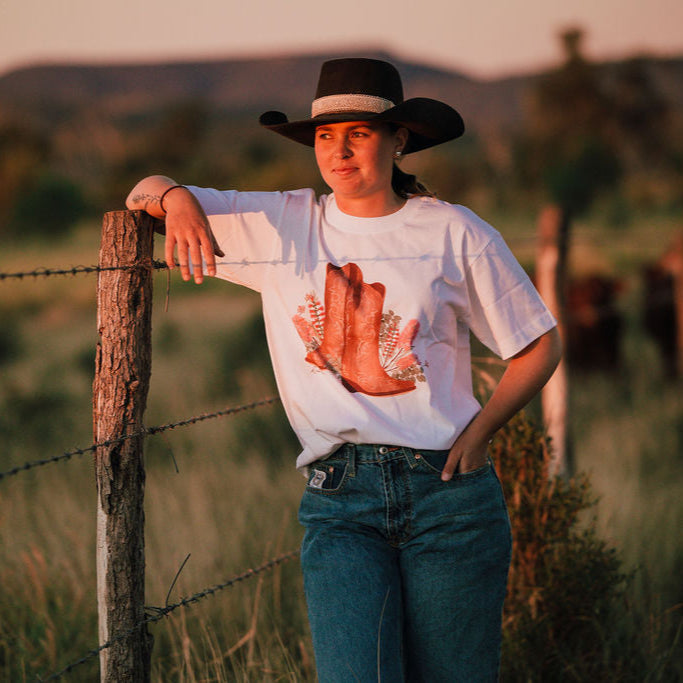 Person wearing a cowboy hat and white t-shirt with a graphic design, standing by a barbed wire fence in a field.
