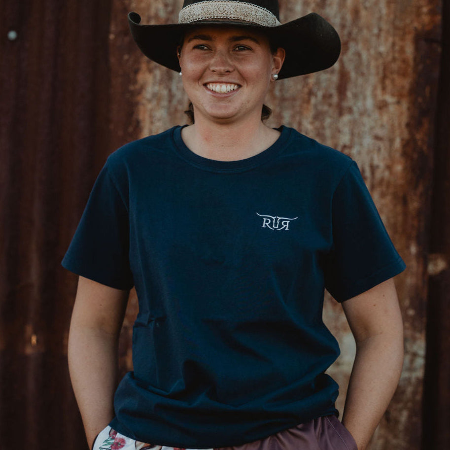 Person wearing a navy blue t-shirt with a logo, patterned shorts, and a wide-brimmed hat against a rusty metal wall.