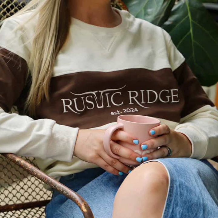 Person wearing a 'Rustic Ridge' sweatshirt holding a pink mug outdoors.