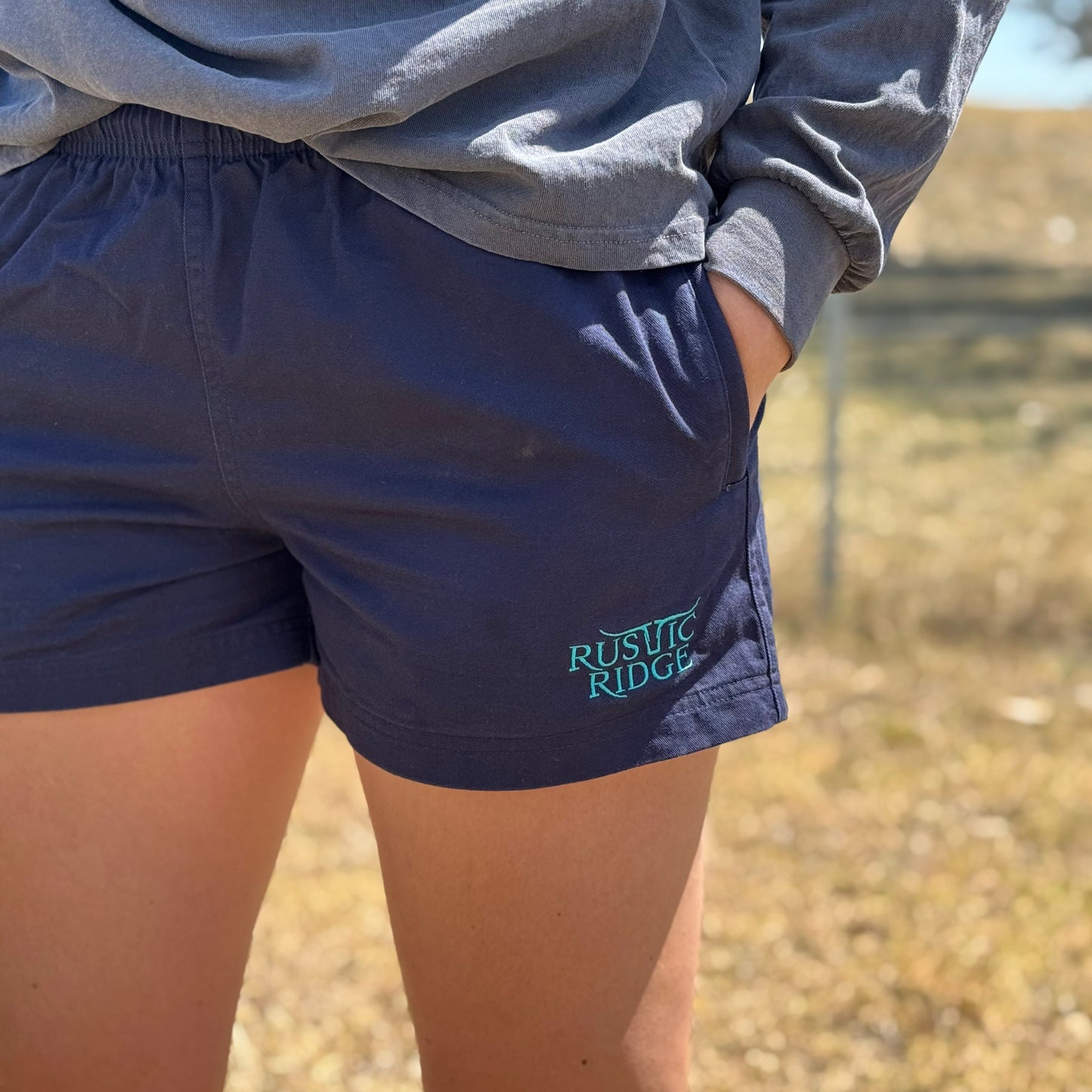 A pair of navy blue work shorts with a visible Rustic Ridge logo on the left leg, designed for unisex wear.
