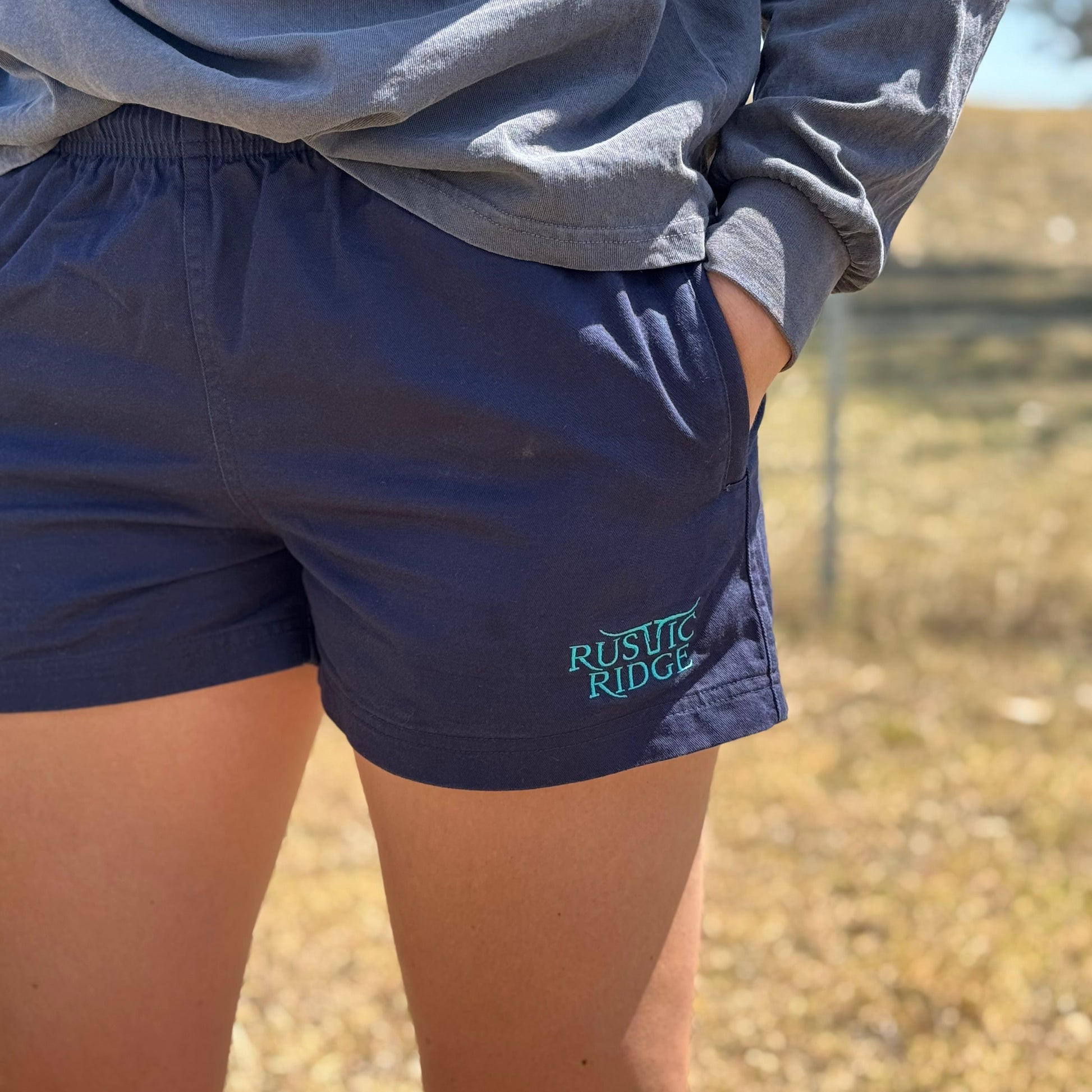 A pair of navy blue work shorts with a visible Rustic Ridge logo on the left leg, designed for unisex wear.