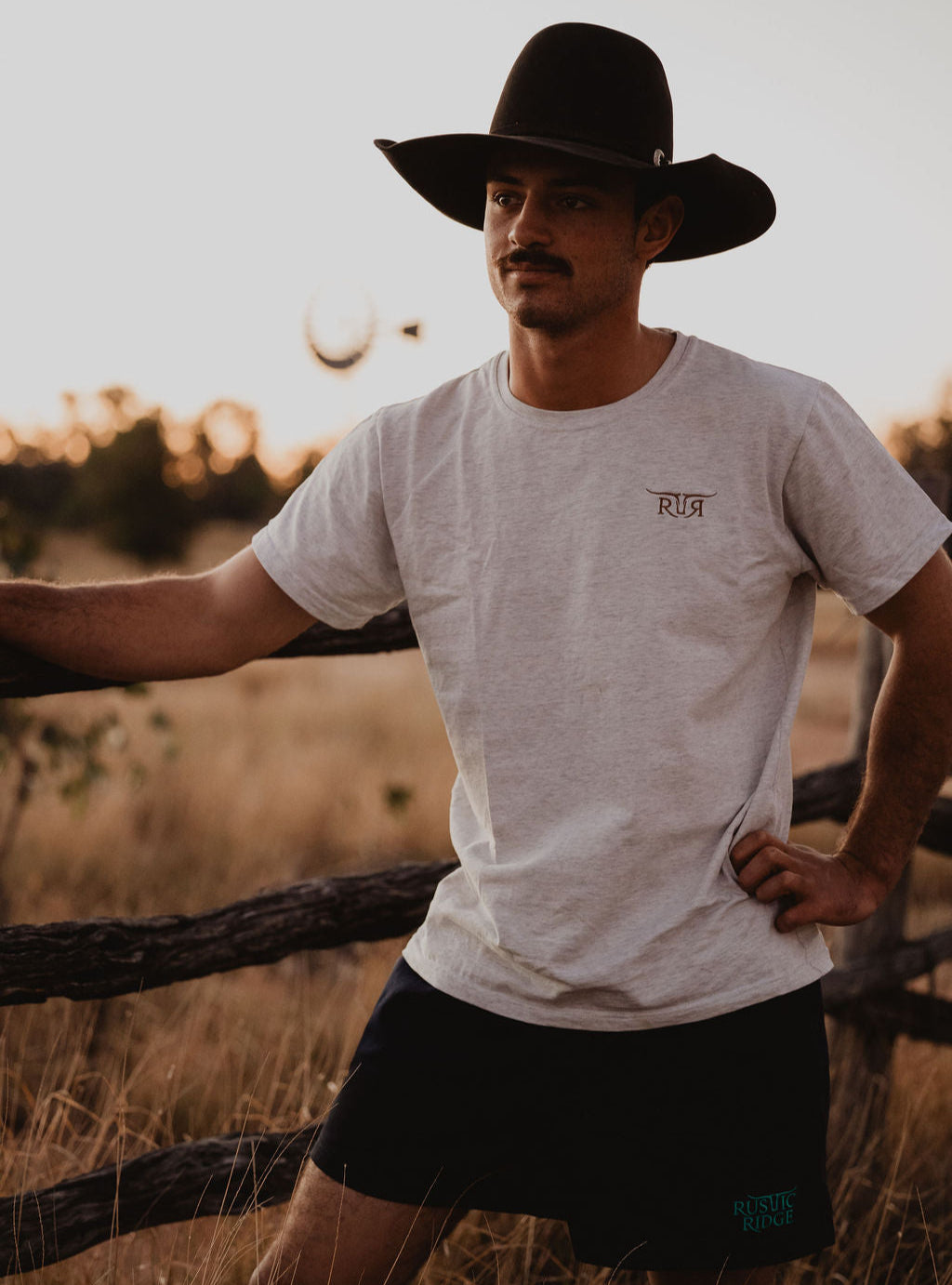 Man wearing a cowboy hat and casual clothing standing in a field with a sunset or sunrise in the background.