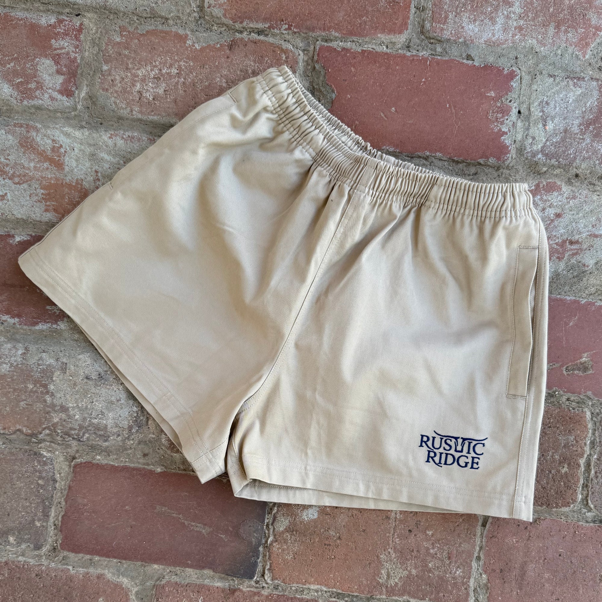 Beige shorts with 'Rustic Ridge' logo on a brick surface
