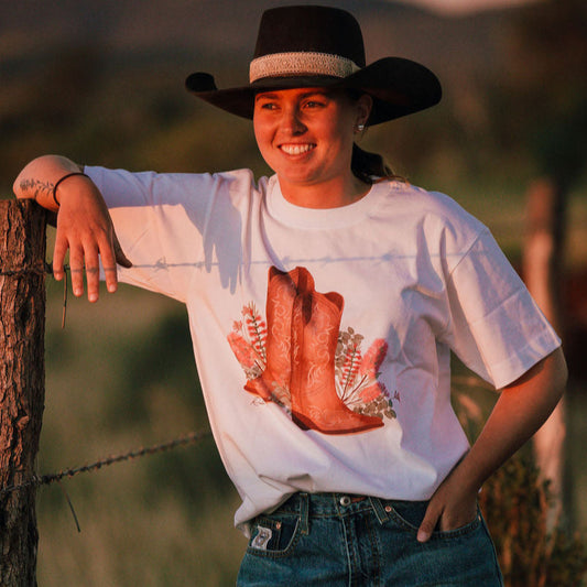 Person wearing a white t-shirt with a red graphic and blue jeans, leaning against a wooden post in a field.