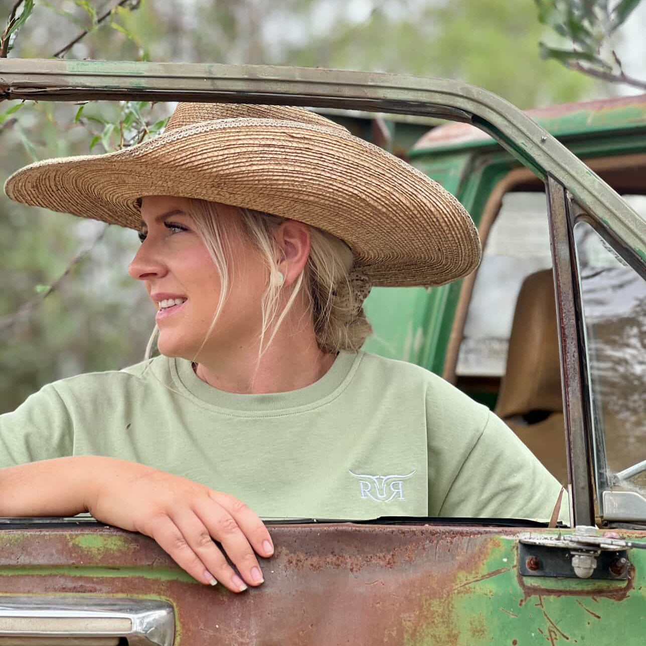 Person wearing a wide-brimmed hat and green shirt leaning on a vehicle outdoors.