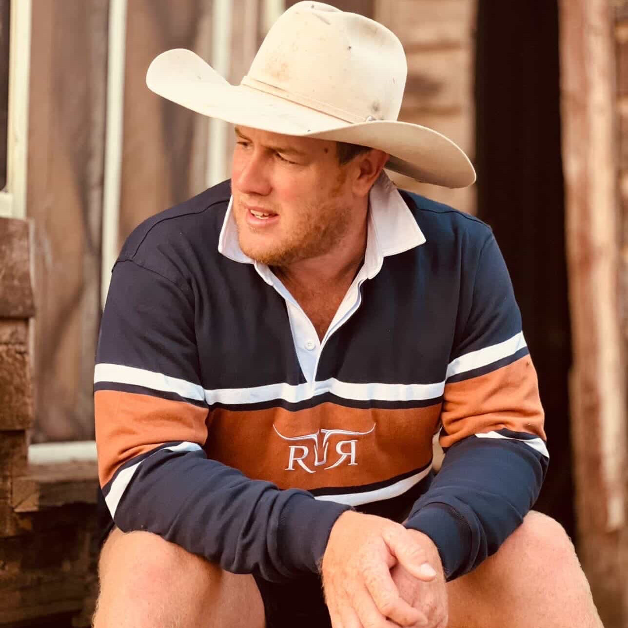 Man wearing a cowboy hat and a rugby jersey with a brand logo, sitting outdoors.