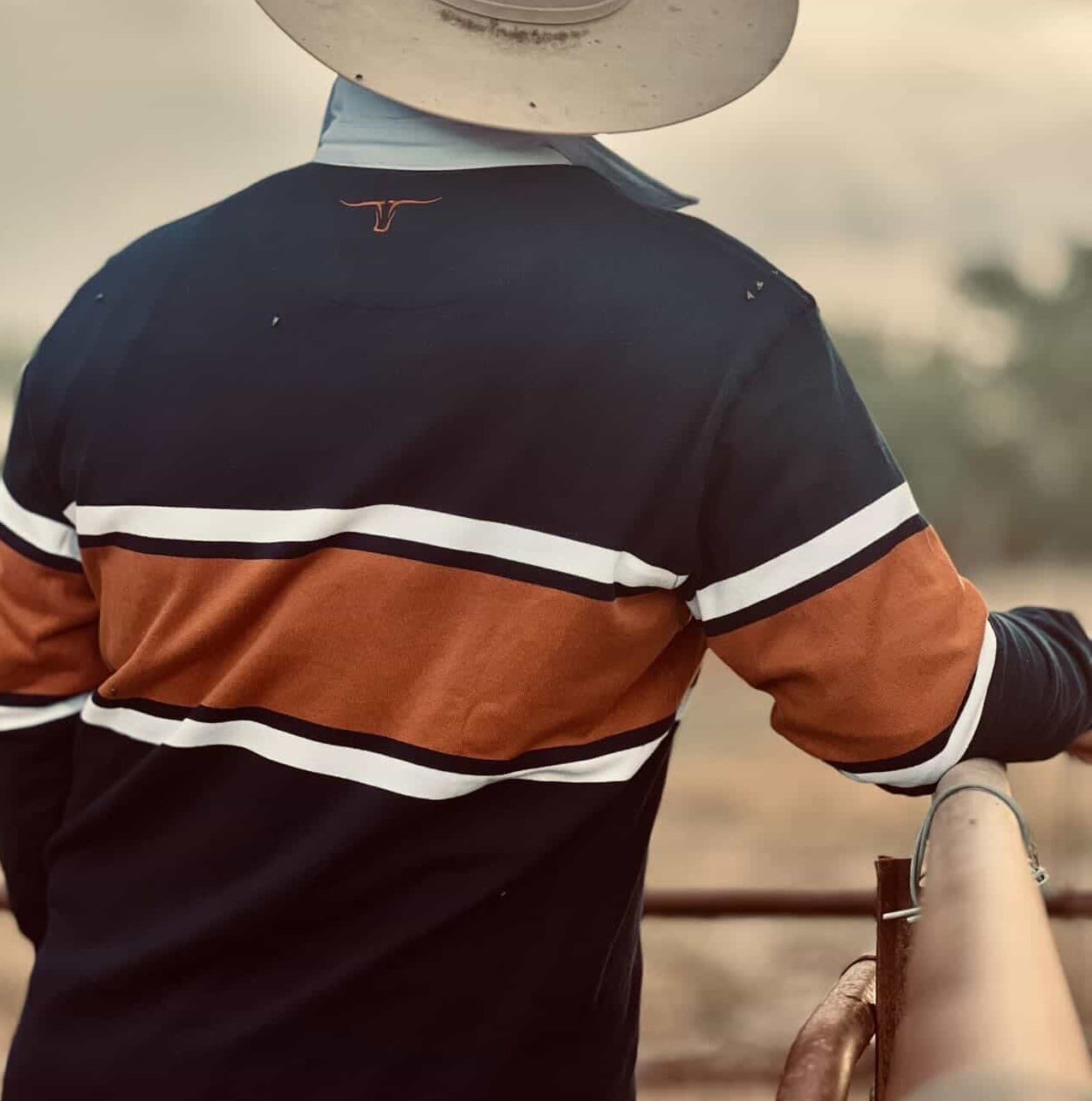 Person wearing a striped shirt and cowboy hat, standing on a fence with a blurred natural background
