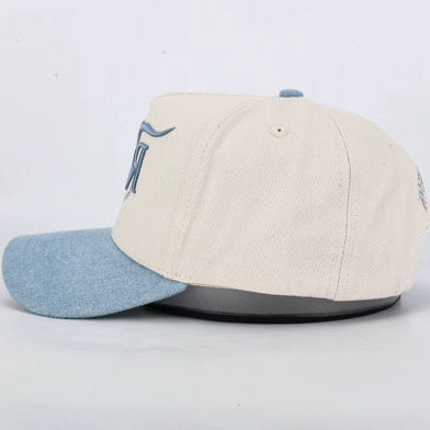 A baseball cap with an off-white crown and a light blue denim-look visor, featuring a blue embroidered logo on the side, displayed on a white background.