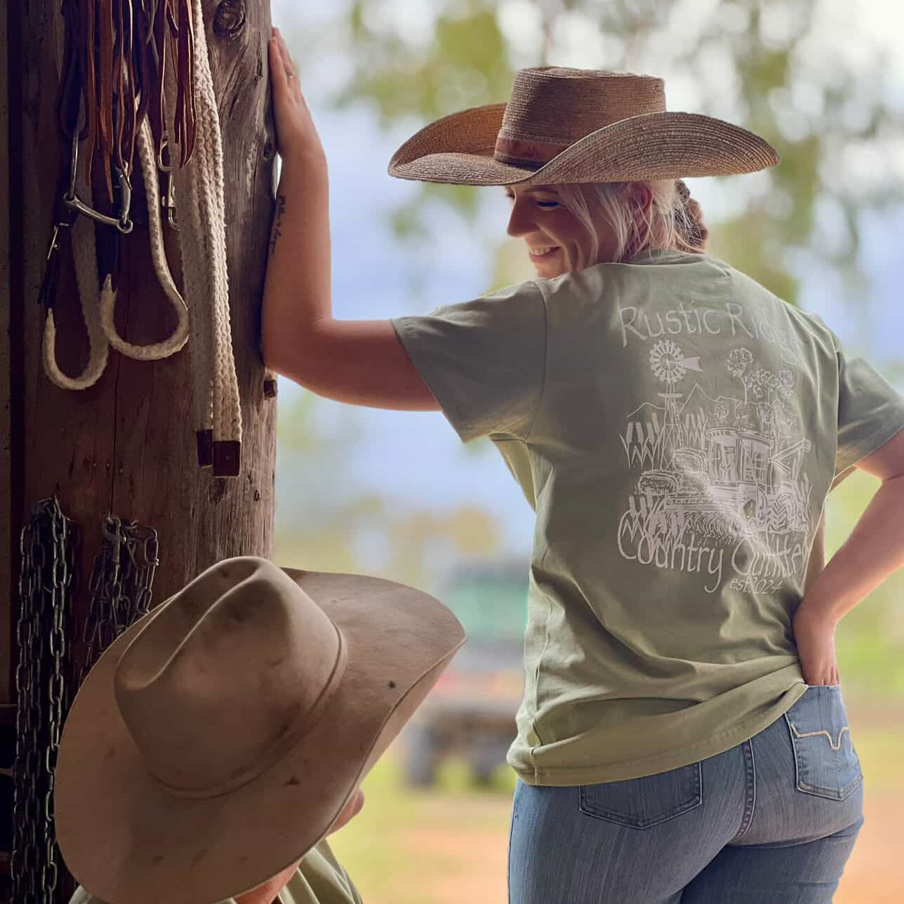 Person wearing a cowboy hat and green t-shirt with a graphic design, standing next to a wooden post with a hat on it.
