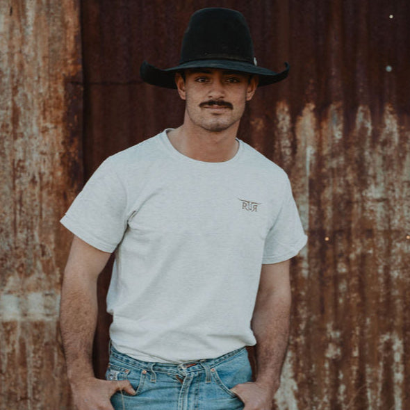 Man wearing a cowboy hat and white t-shirt standing in front of a rusty corrugated metal wall.