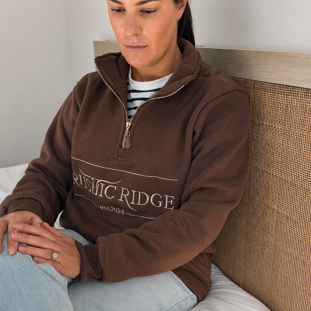 A woman sits on a bed wearing a brown quarter-zip sweatshirt with 'RUSTIC RIDGE est.2024' printed across the chest, paired with light blue jeans and a striped shirt.