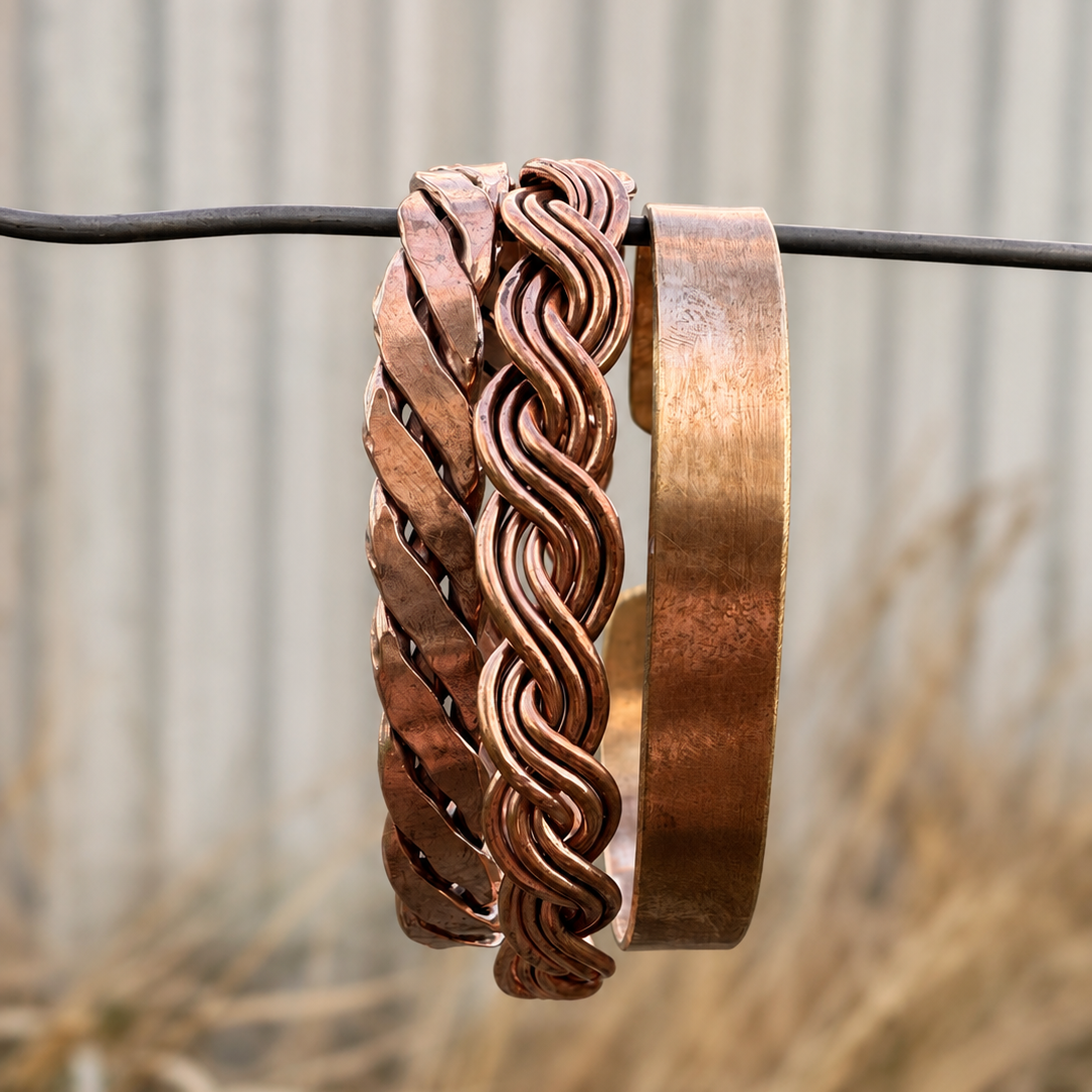 Three copper-toned metal bangles, one with a woven braided pattern and one smooth, displayed side-by-side on a black wire against a blurred background of dry grass and a corrugated metal wall.