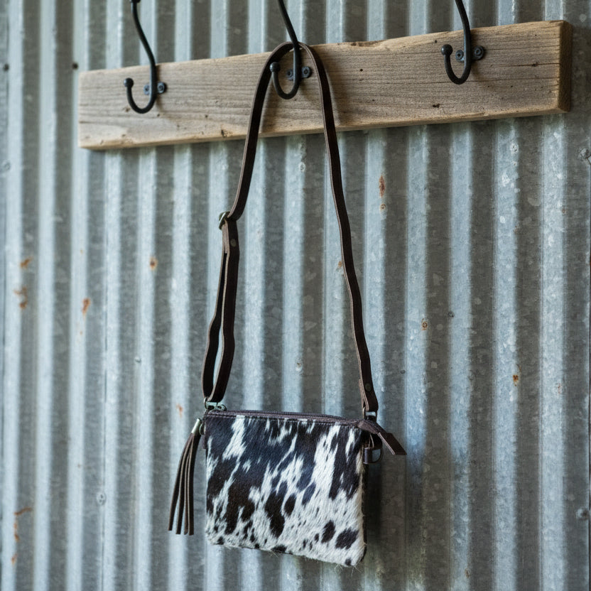 Cow print handbag with brown strap hanging on a wooden rack against a corrugated metal wall.