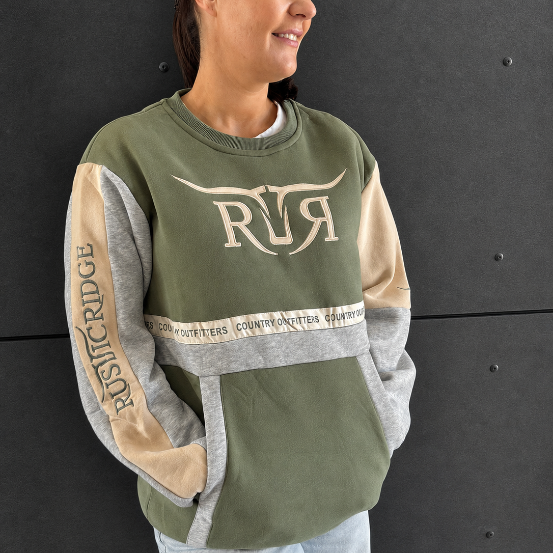 A woman wearing an olive green, gray, and beige color-blocked sweatshirt with 'RR' and 'Rustic ridge' text graphics, paired with light blue jeans, standing against a dark gray wall.