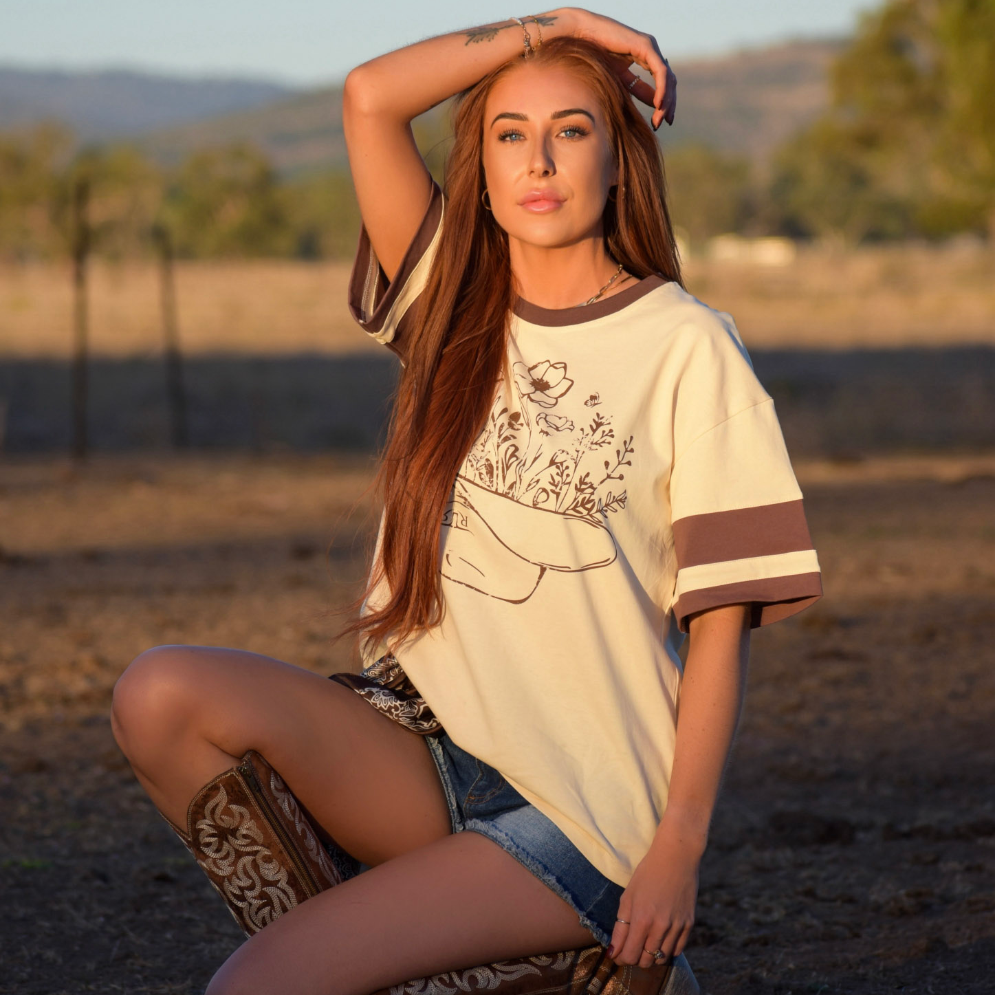 Woman in a beige top and denim shorts kneeling on a dirt ground with trees and mountains in the background.
