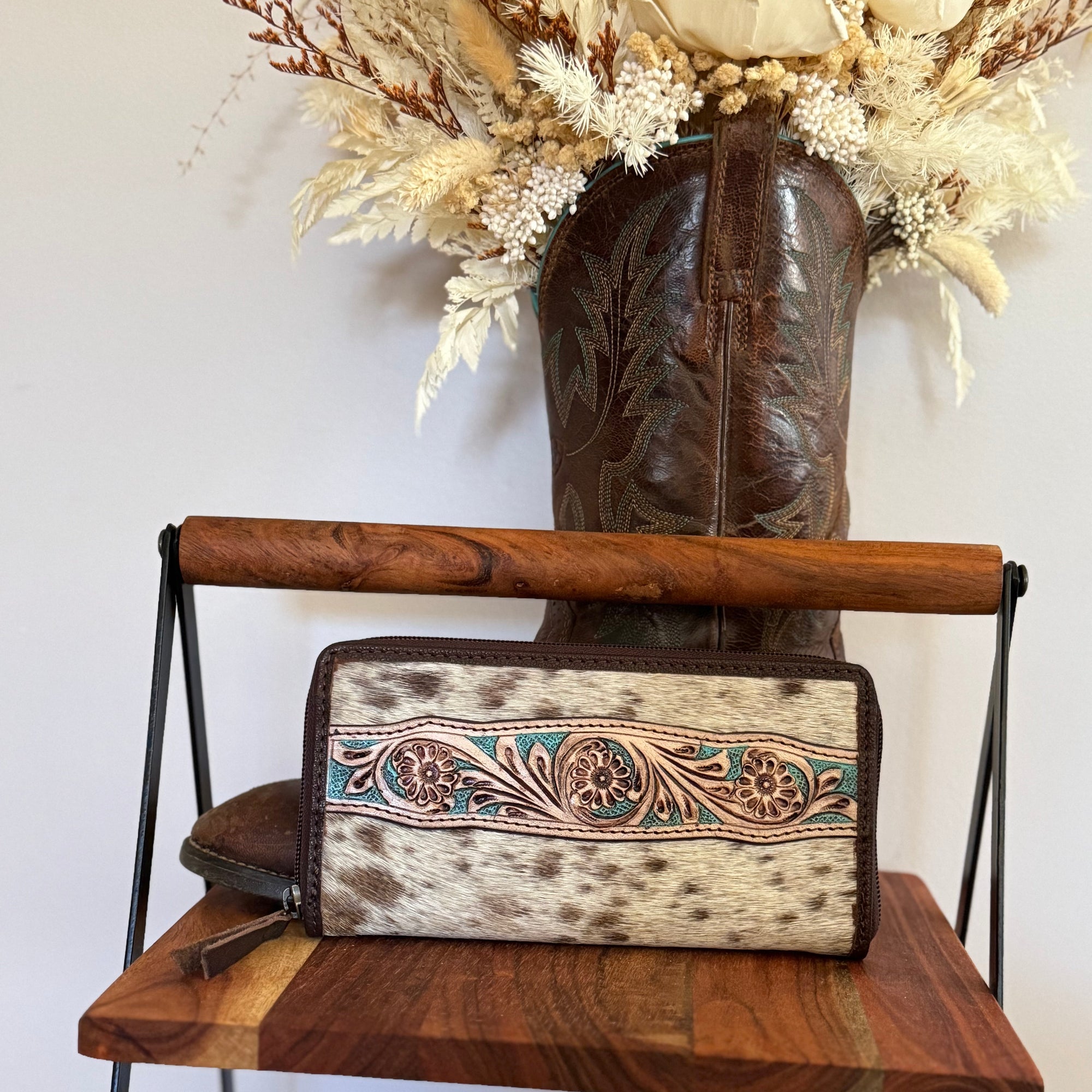 Cowhide leather clutch with floral embroidery on a wooden shelf with a cowboy boot and flowers in the background.