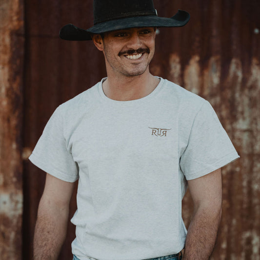 Man wearing a white t-shirt with a logo and blue jeans, standing against a rusty metal wall.