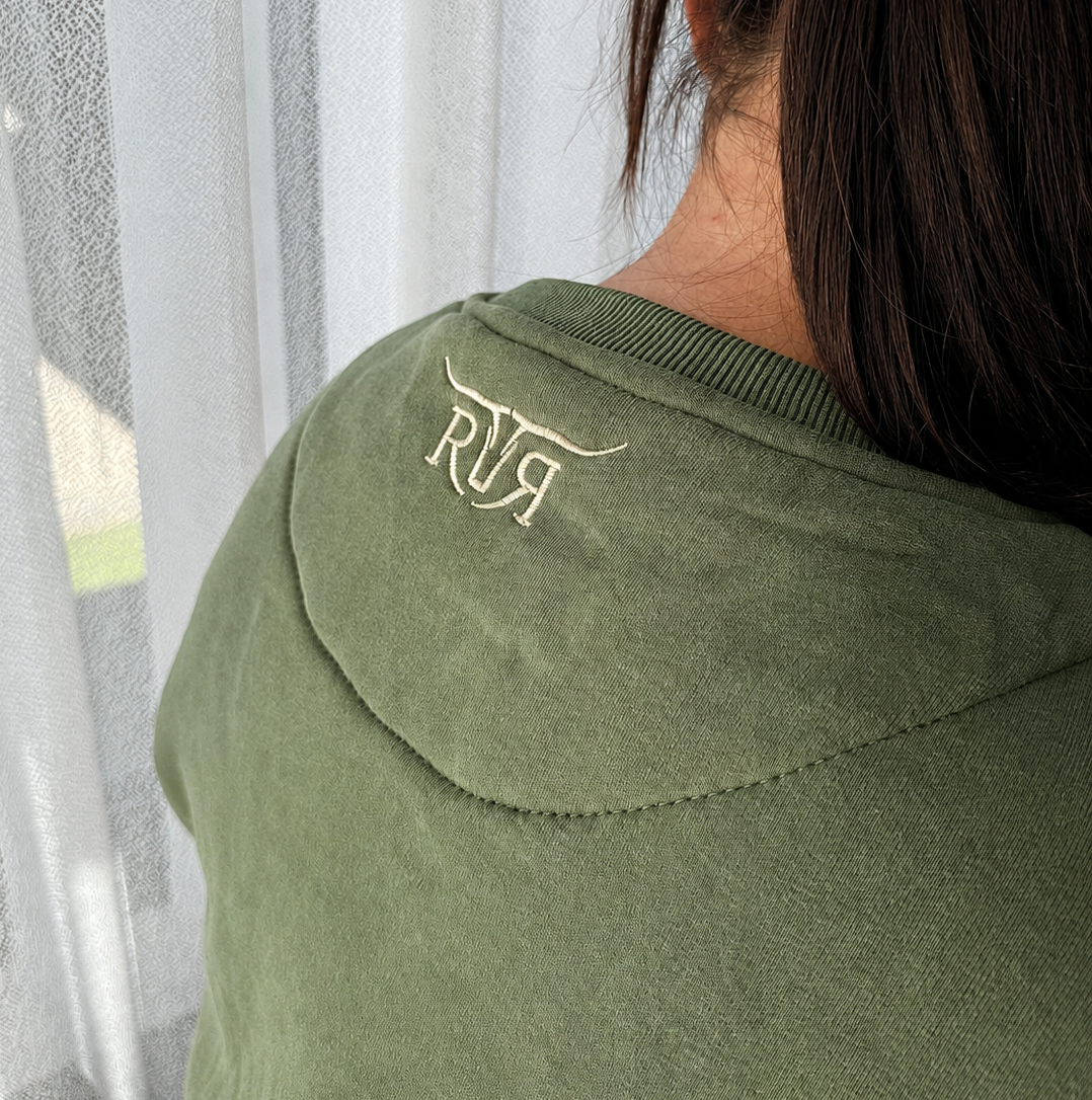 A close-up back view of a person wearing an olive green hooded sweatshirt with a small white embroidered logo on the upper back, standing in front of sheer white curtains.