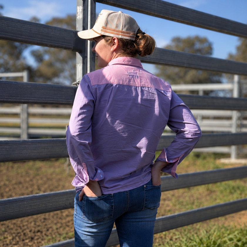 A woman stands outdoors in front of a wooden fence, viewed from behind. She is wearing a lavender-purple long-sleeve button-up shirt, blue jeans, and a beige baseball cap.
