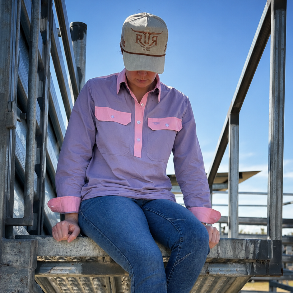 A person sitting on a metal ranch gate outdoors, wearing a lavender long-sleeve western-style shirt with pink pocket flaps and pink piping, blue jeans, brown boots, and a beige cap with 'RUG' embroidered on the front.