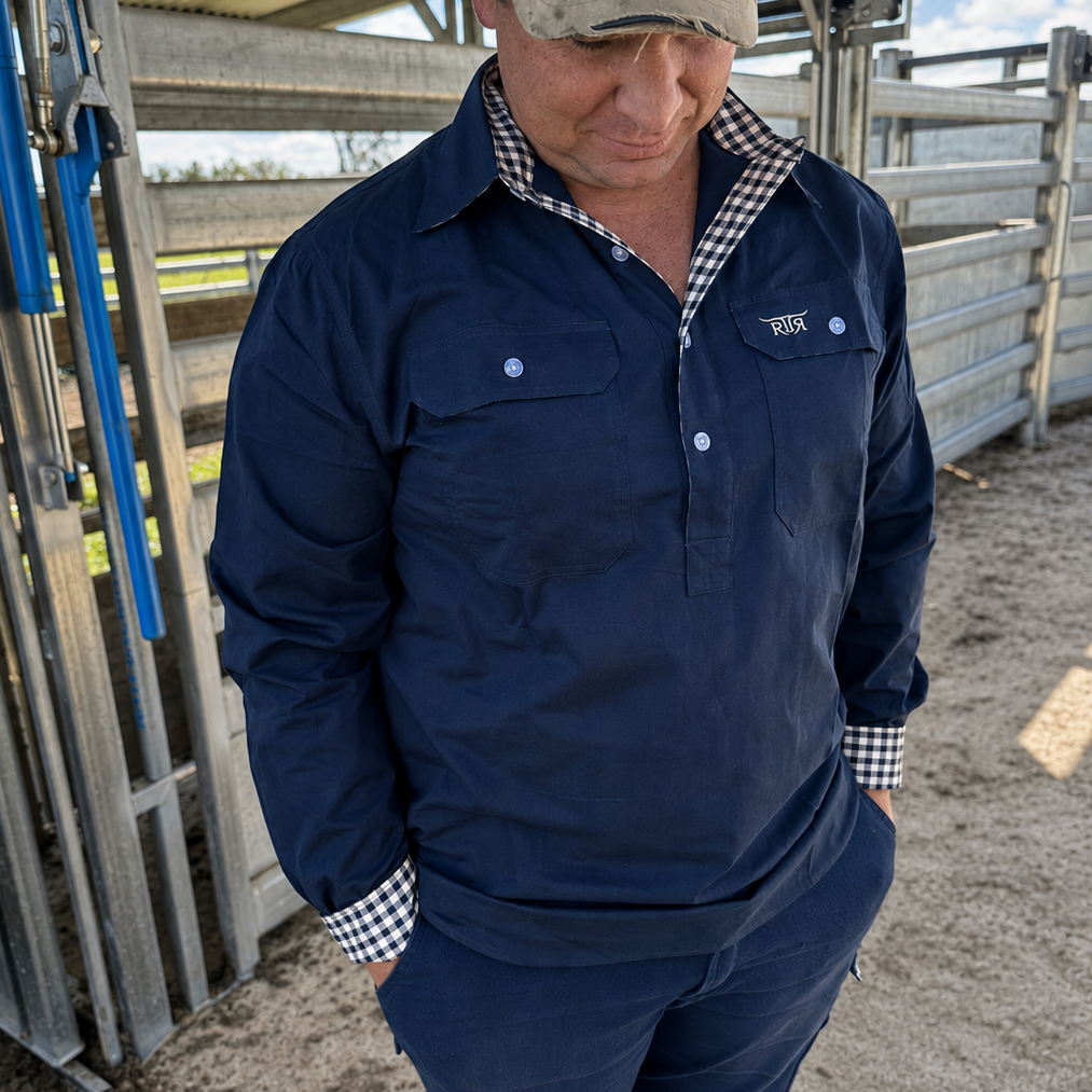 A man wearing a navy blue farm work shirt with a small white embroidered logo on the left chest, standing in an outdoor barn or livestock pen.