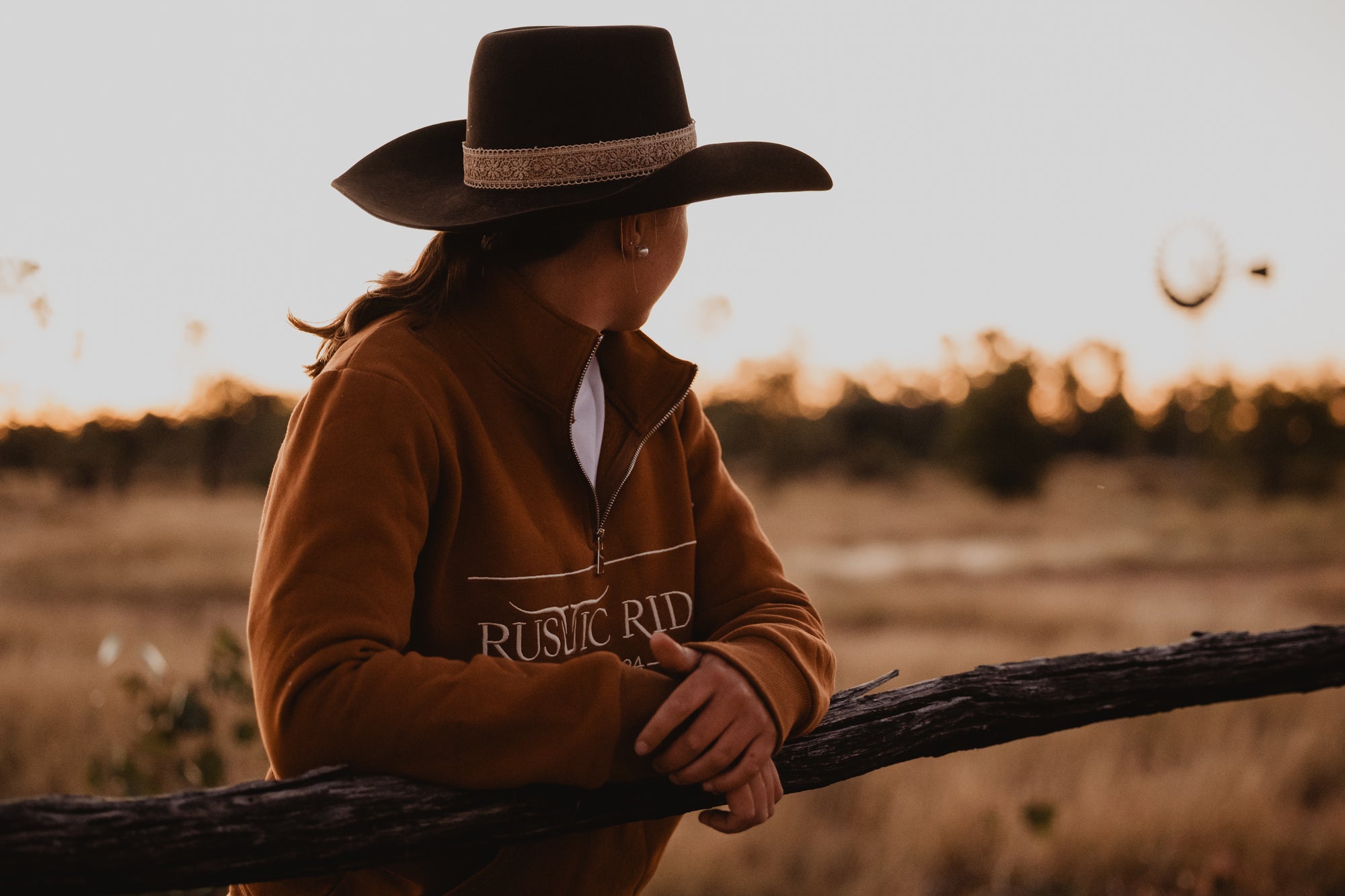 Person wearing a cowboy hat and brown jacket with 'Rustic Ridge' logo in a field at sunset.