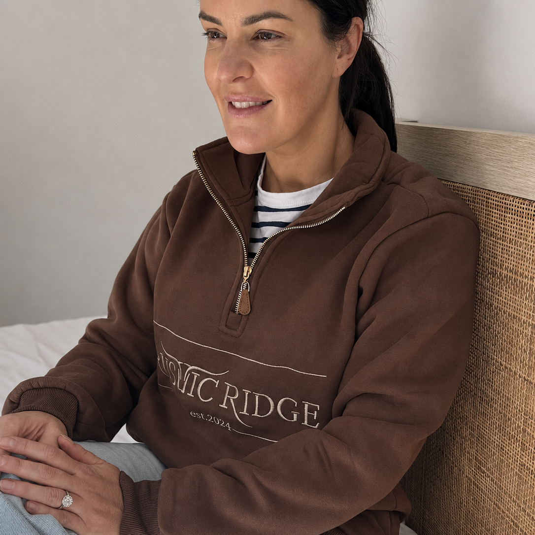 A woman sitting on a bed wearing a brown quarter-zip sweatshirt with 'RUSTIC RIDGE' printed across the chest, paired with a striped shirt and light blue jeans.