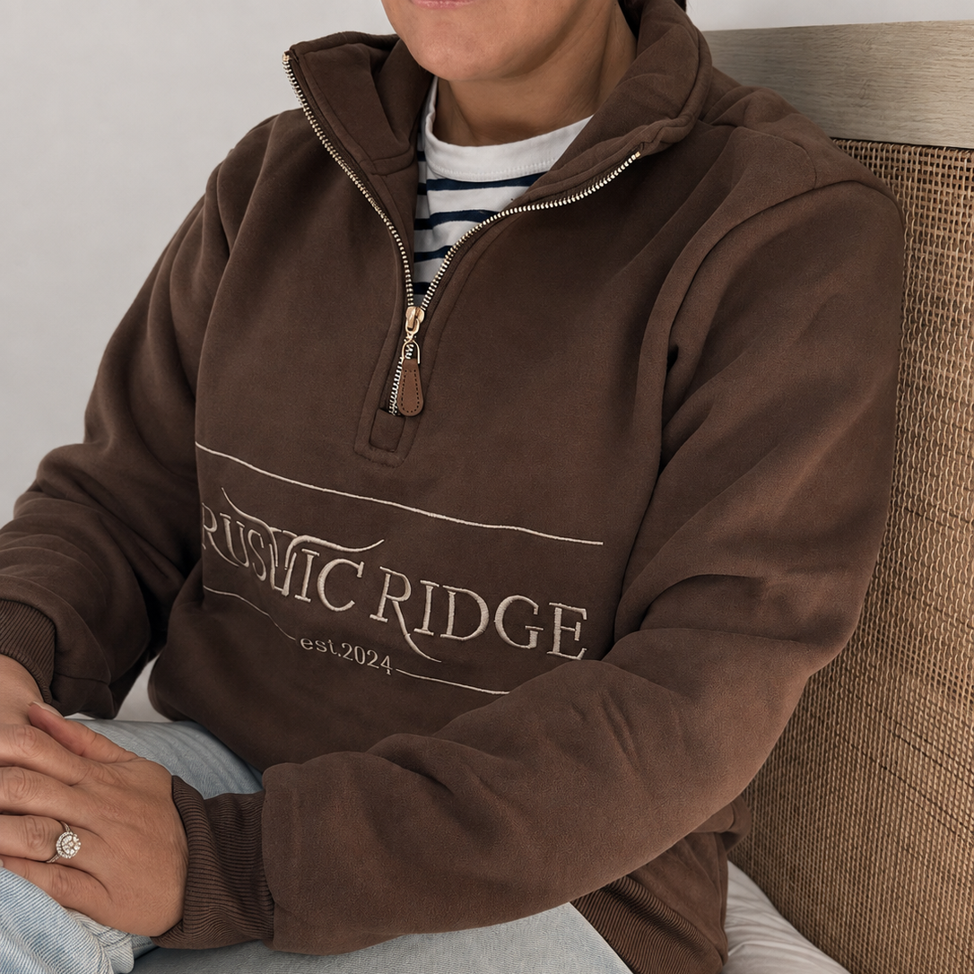 A person wearing a brown quarter-zip sweatshirt with 'RUSTIC RIDGE' printed across the chest, paired with a striped shirt and light blue jeans, sitting on a bed.