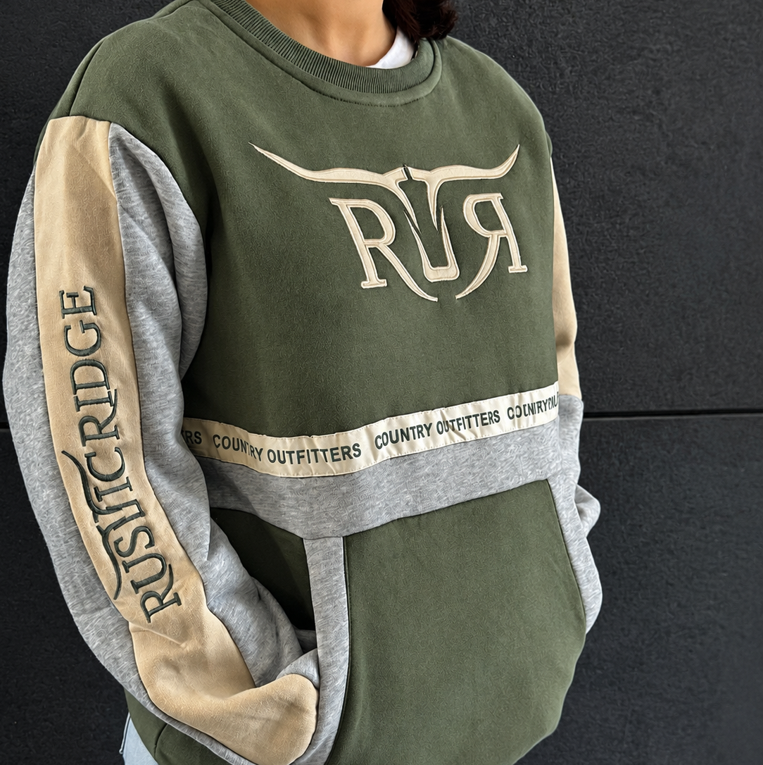 A person wearing an olive green and heather gray colorblock sweatshirt with beige panels and a large 'RR' logo on the chest, paired with light blue jeans against a dark gray wall.