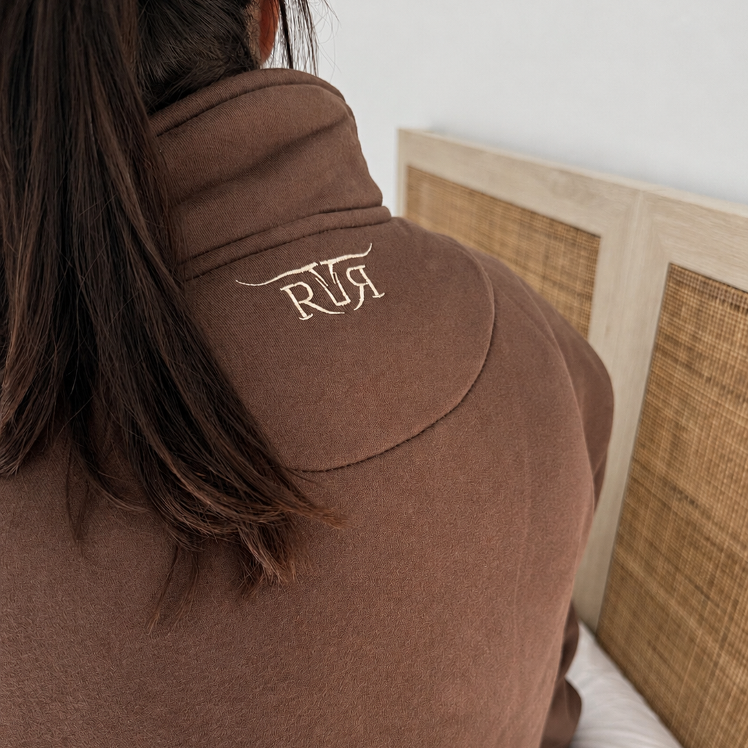 A person wearing a brown fleece pullover sweatshirt with a high neck and a white embroidered logo on the back, sitting on a bed with a woven headboard.