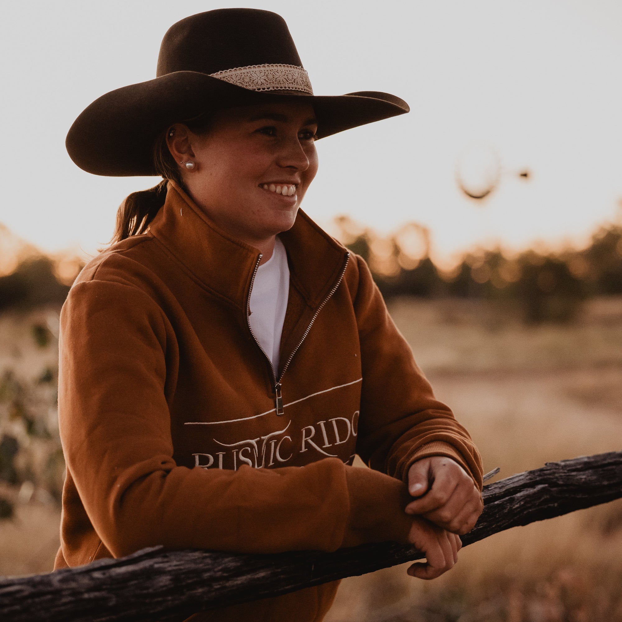 Woman in a brown jacket and cowboy hat standing in a field with a sunset.