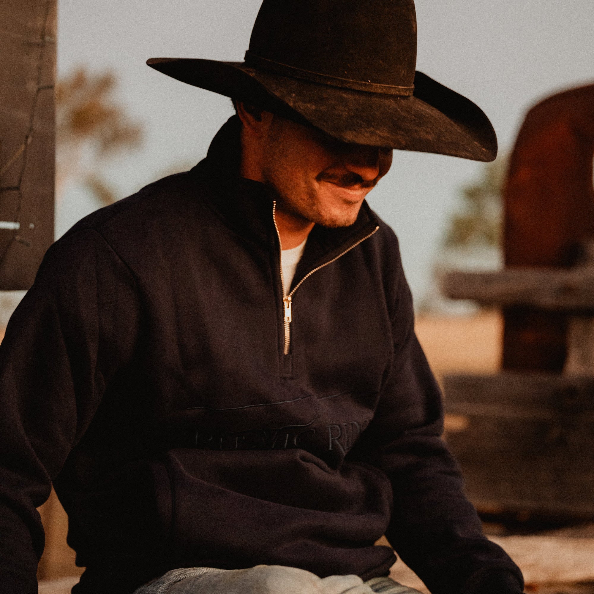 Man wearing a cowboy hat and dark jacket sitting outdoors.