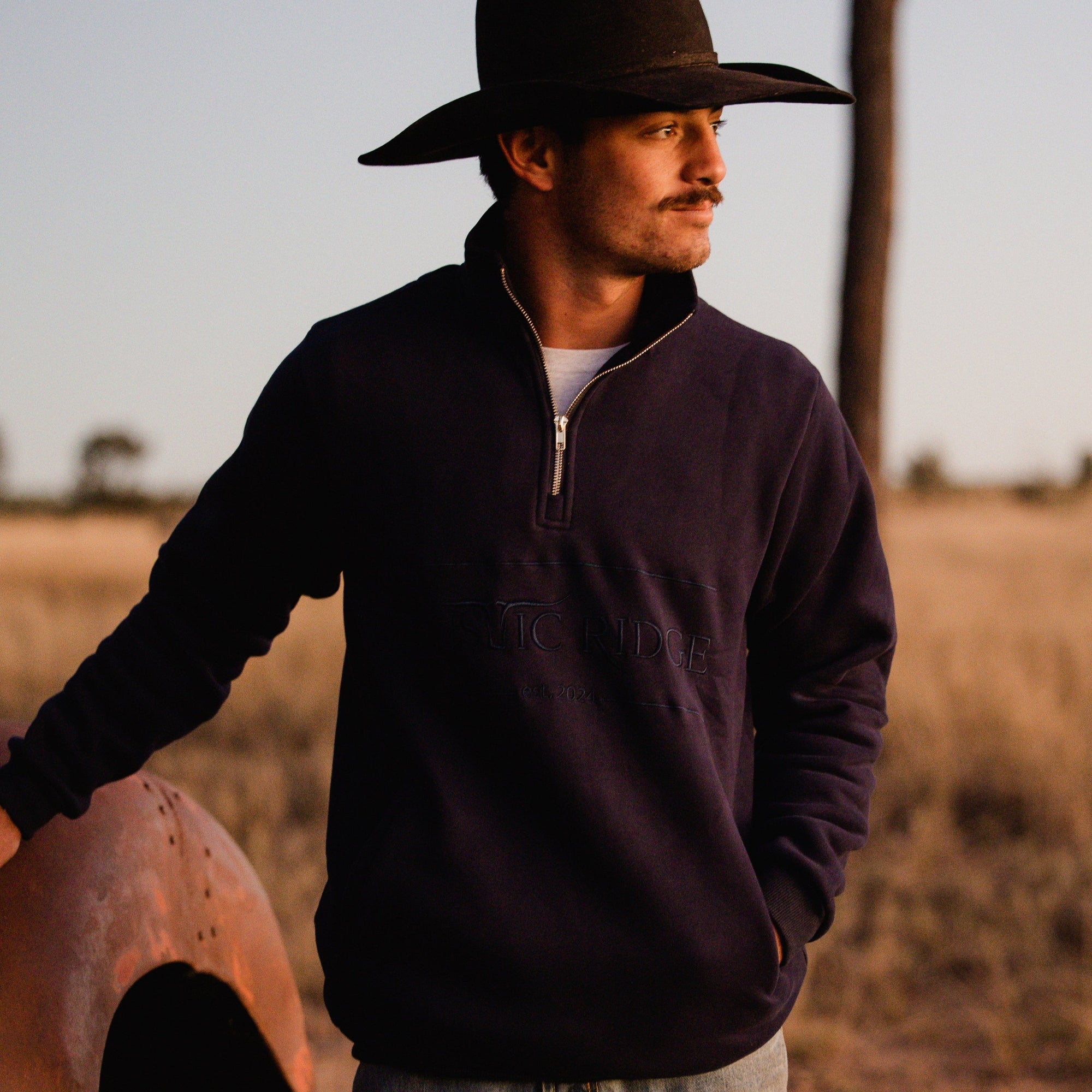 Man wearing a cowboy hat and dark jacket standing next to an old vehicle in a field.
