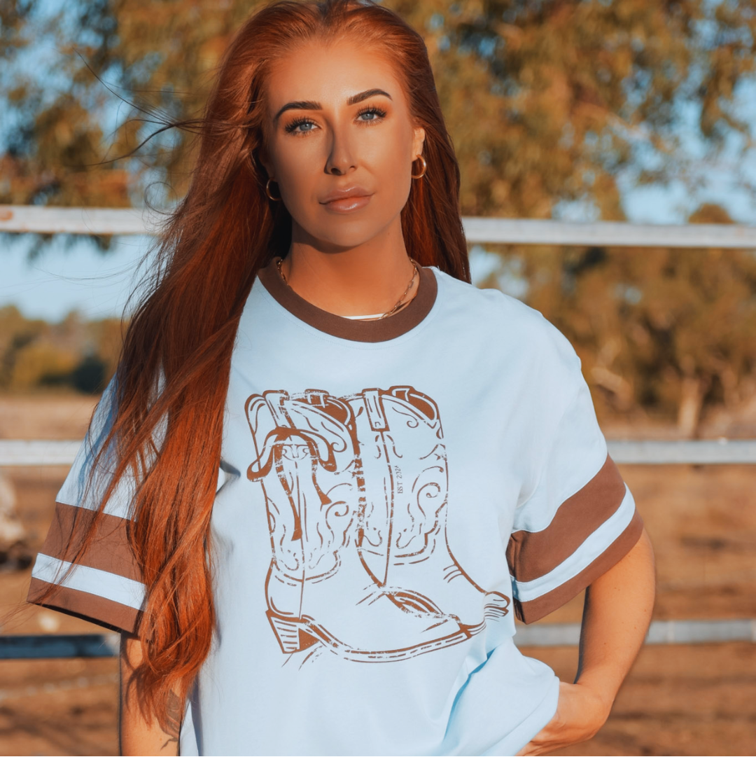 Woman wearing a white t-shirt with a graphic of cowboy boots outdoors.