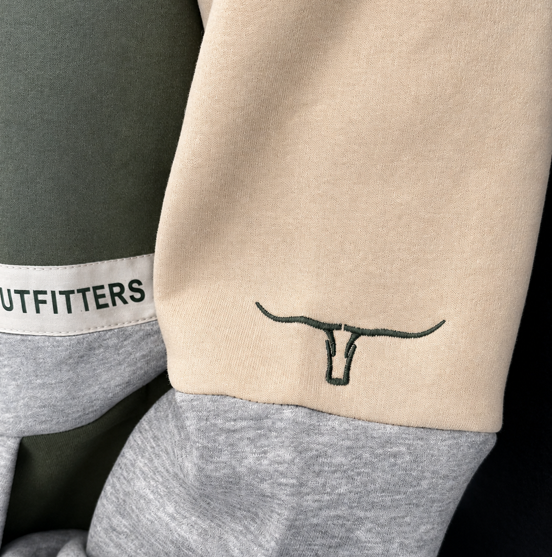 Close-up of a colorblock sweatshirt sleeve featuring beige, gray, and dark olive green sections, with a black embroidered longhorn logo on the beige cuff and a white brand label reading COUNTRY OUTFITTERS on the gray cuff.