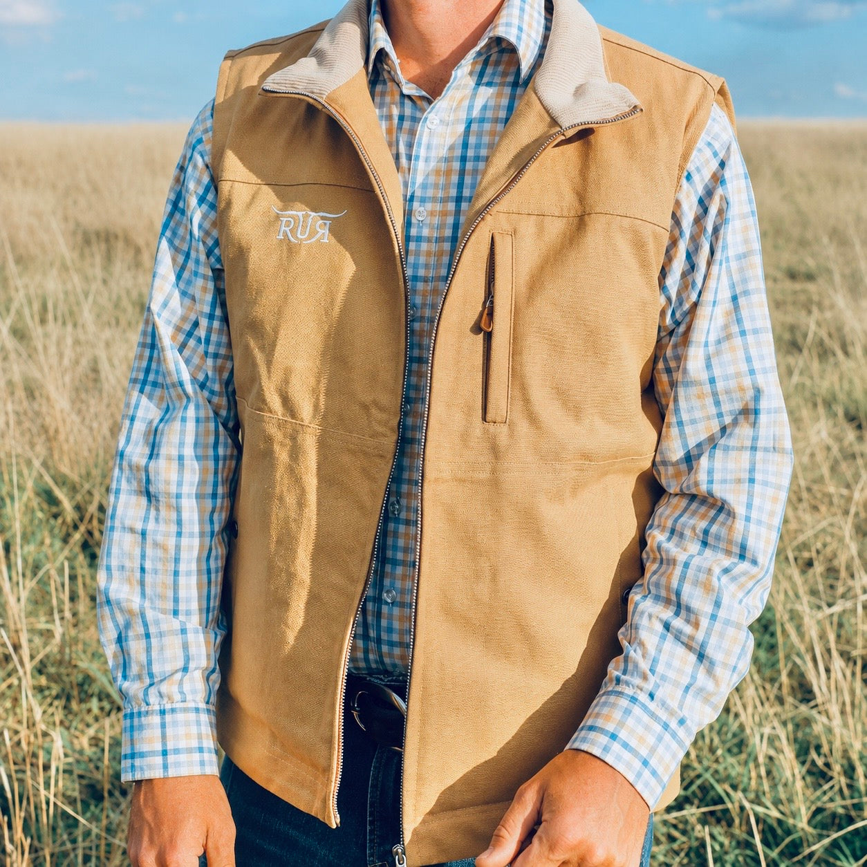 A man wearing a tan corduroy vest with a light gray fleece collar over a blue and white plaid button-down shirt, standing in a grassy field.