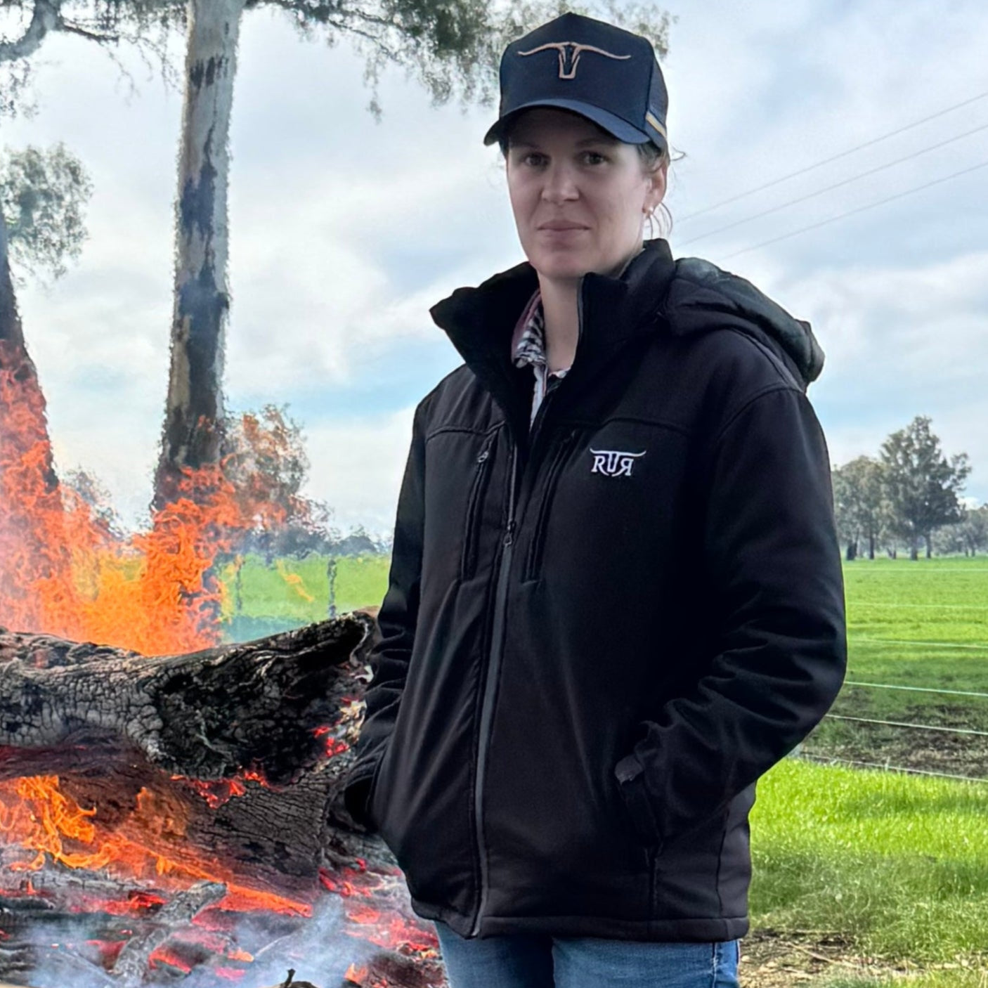 Person standing near a fire in a field with trees and power lines in the background