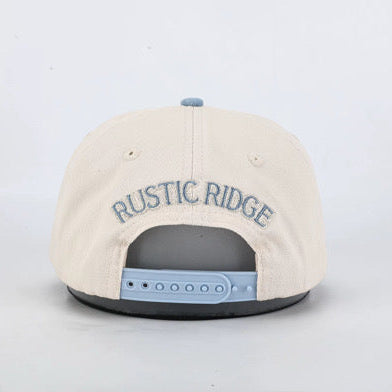 The back of a cream-colored snapback cap featuring 'RUSTIC RIDGE' embroidered in blue across the crown, with a light blue plastic snapback closure and a black undercap, set against a plain light gray background.