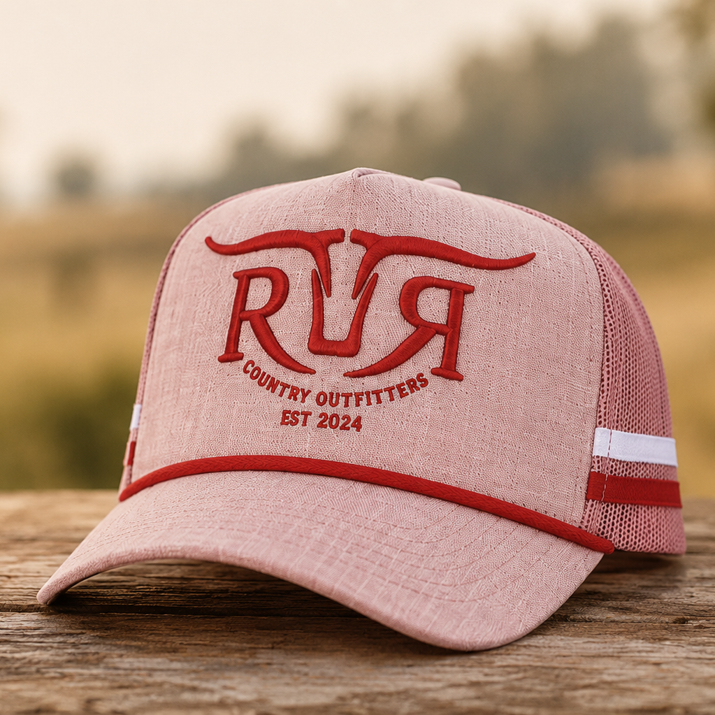 A dusty pink trucker hat with red embroidered lettering reading 'RR COUNTRY OUTFITTERS EST 2024' sits on a wooden surface with a blurred outdoor background.