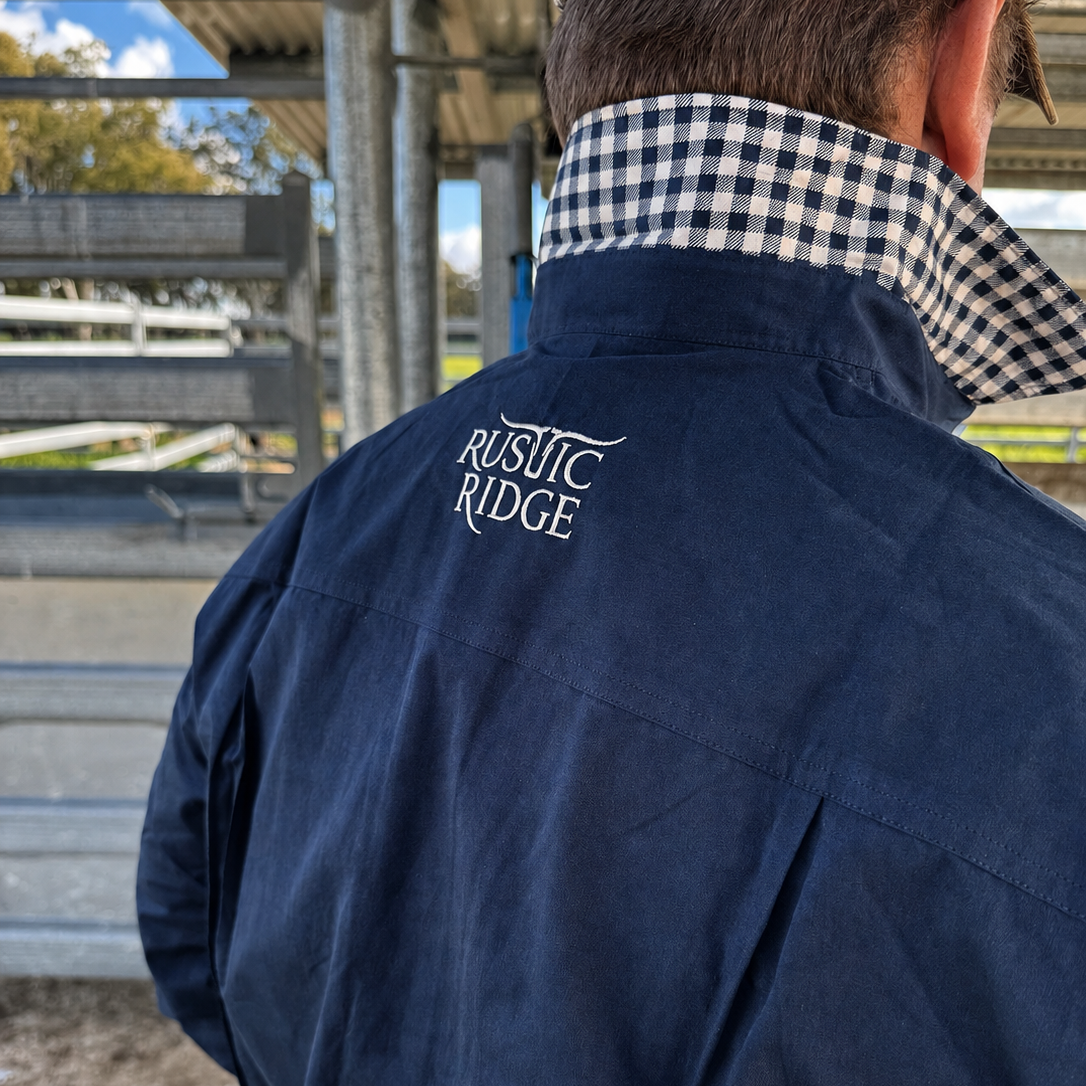 A man viewed from behind wearing a navy blue shirt jacket with 'RUSITIC RIDGE' embroidered on the back, featuring a blue and white gingham collar and a tan cap in an outdoor cattle yard setting.