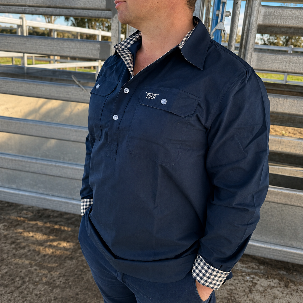 A man wearing a navy blue long-sleeve button-up shirt, matching navy pants, a brown trucker hat, and a checkered shirt collar, standing in an outdoor cattle yards. 