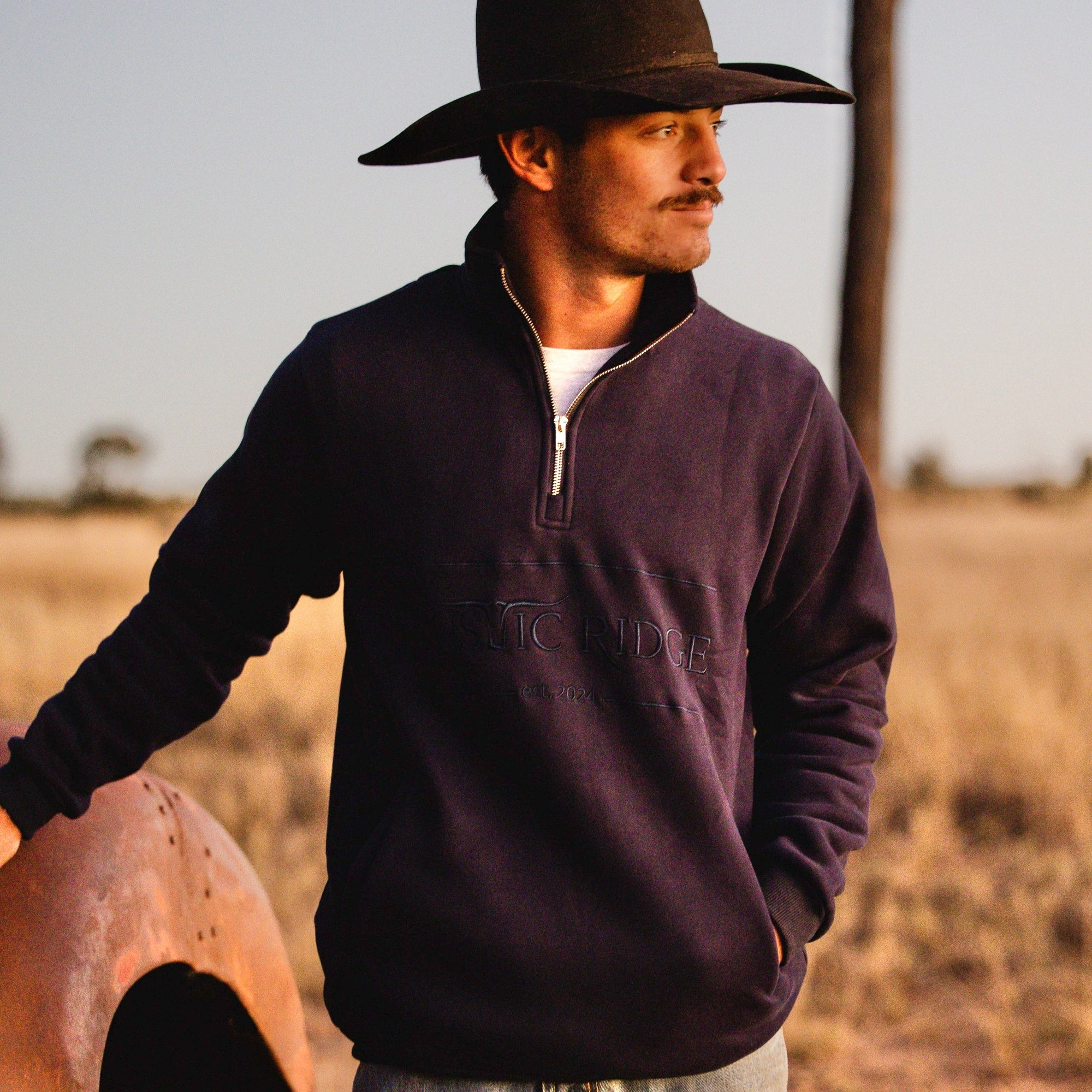 Man wearing a dark sweater and cowboy hat standing in a field.