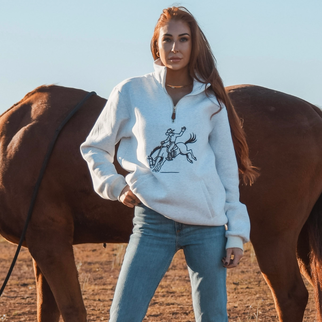 Woman wearing a white hoodie with a horse graphic, standing next to a horse in an open field.