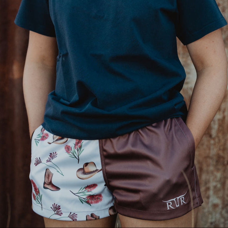 Person wearing a navy blue t-shirt with a logo, floral shorts, and maroon shorts in front of a rusty metal wall.